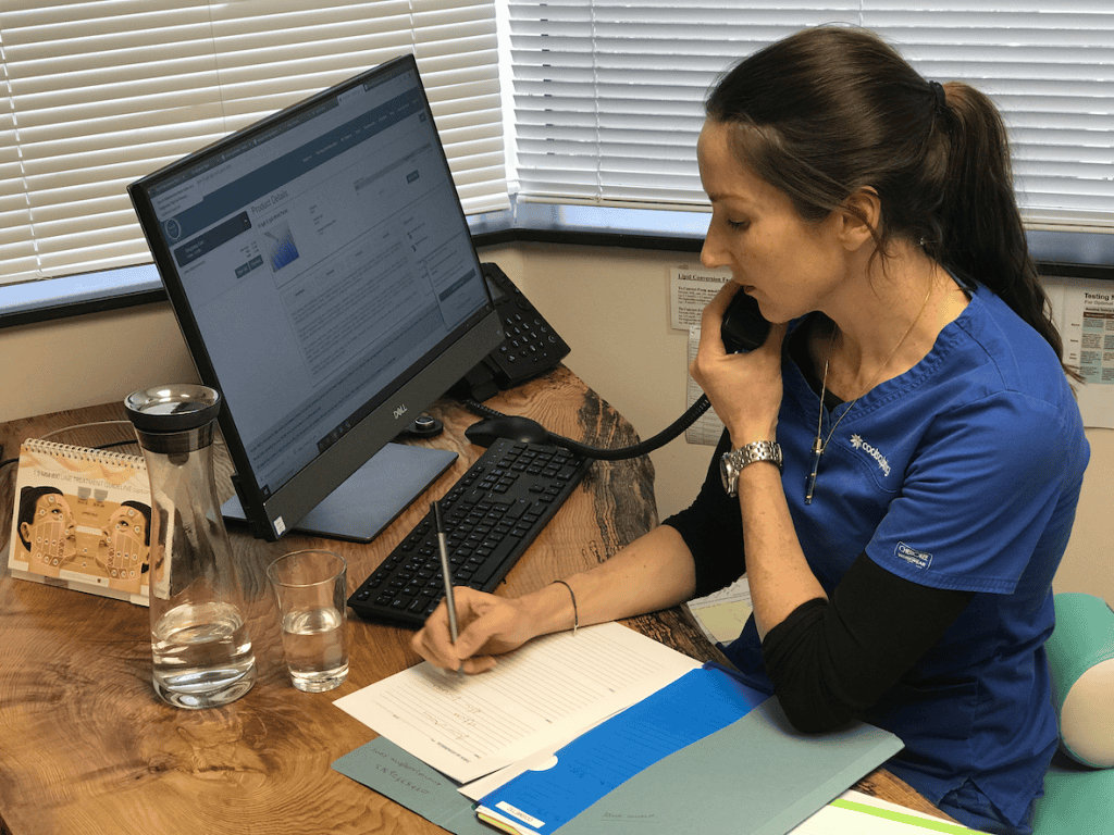 Annelize Meyer in a blue uniform is speaking on the phone while taking notes at a desk with a computer and a glass of water, ensuring online consultations provide a lifeline to new and existing patients at Meyer Clinic. - Meyer Clinic