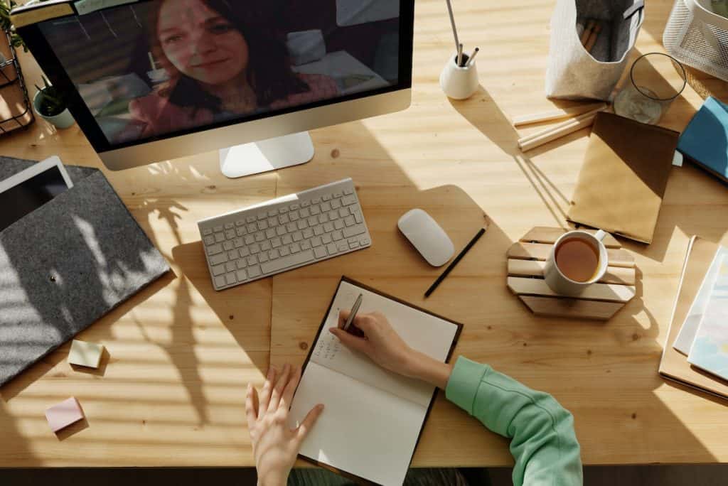 Person taking notes at a wooden desk during a video call on a computer monitor, jotting down tips on staying fit and healthy while working from home. Desk items include a notebook, keyboard, mouse, coffee cup, and folders. - Meyer Clinic