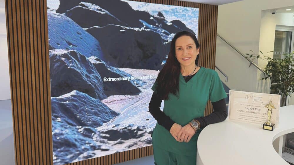 A woman in green scrubs stands at a counter with a MiA Award. Behind her, a large screen displays an image of mountains and text: Extraordinary health and beauty. Her work at the aesthetic clinic focuses on empowering women through menopause and beyond. - Meyer Clinic