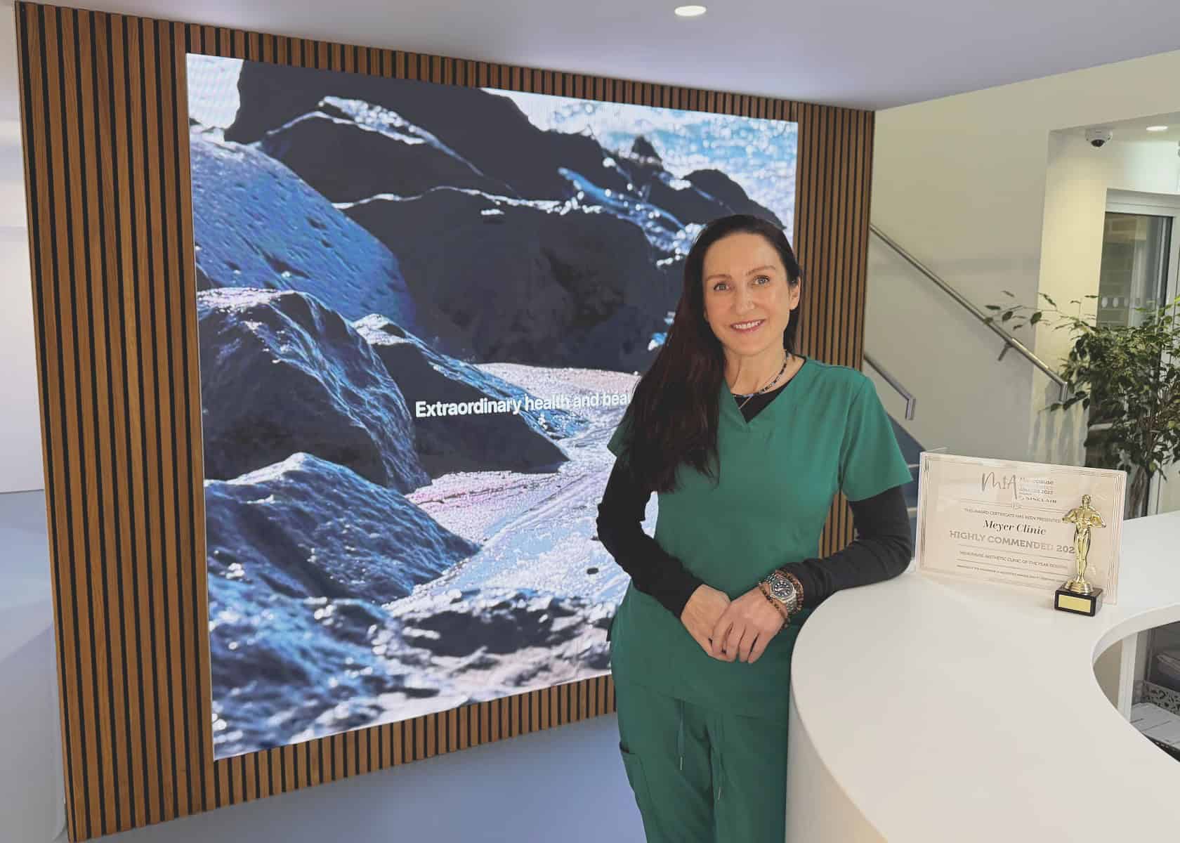 A woman in green scrubs stands at a counter with a MiA Award. Behind her, a large screen displays an image of mountains and text: Extraordinary health and beauty. Her work at the aesthetic clinic focuses on empowering women through menopause and beyond. - Meyer Clinic